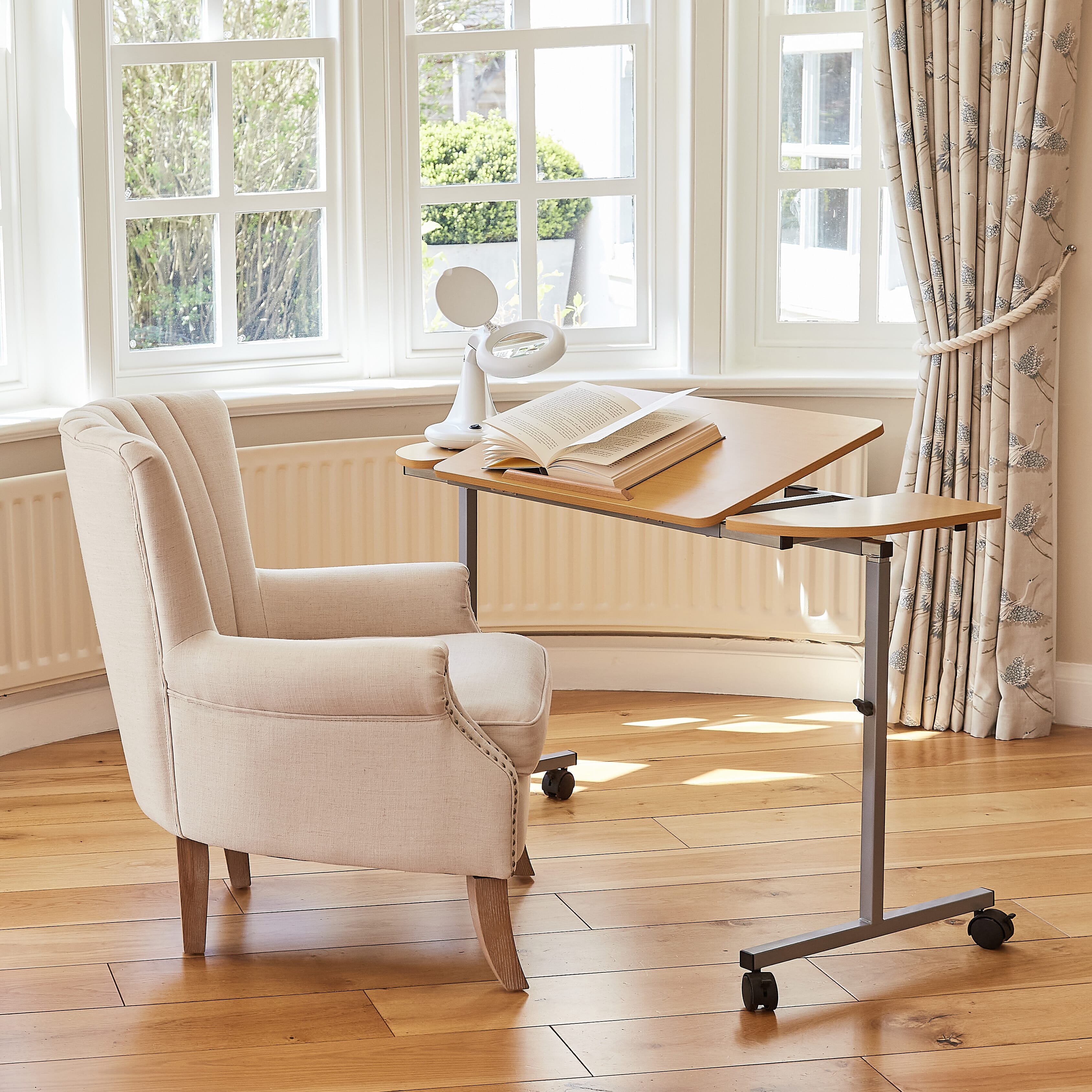 Angled view of NRS Adjustable Overbed Table with beech-effect tilting table top and white U-shaped base on castors against a neutral background.