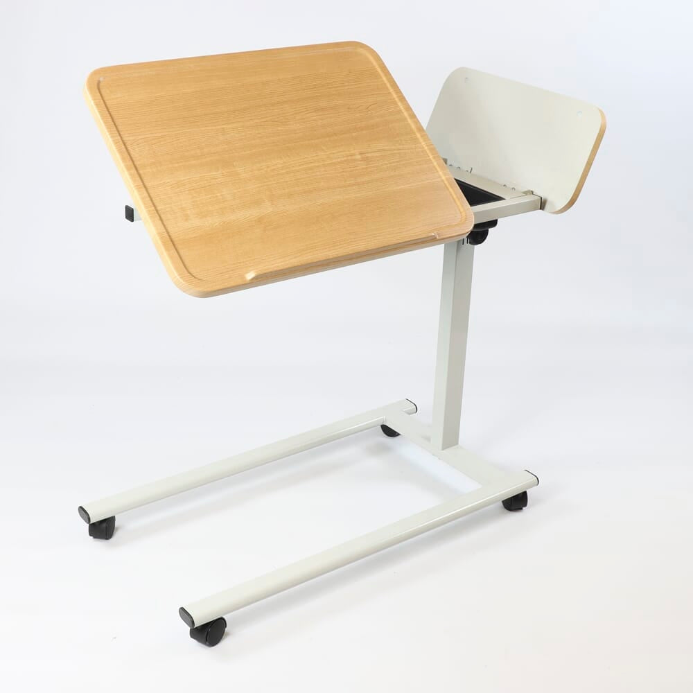 Angled view of NRS Easylift Home Tilting Overbed Table with beech-effect tilting table top and white U-shaped base on castors against a neutral background.