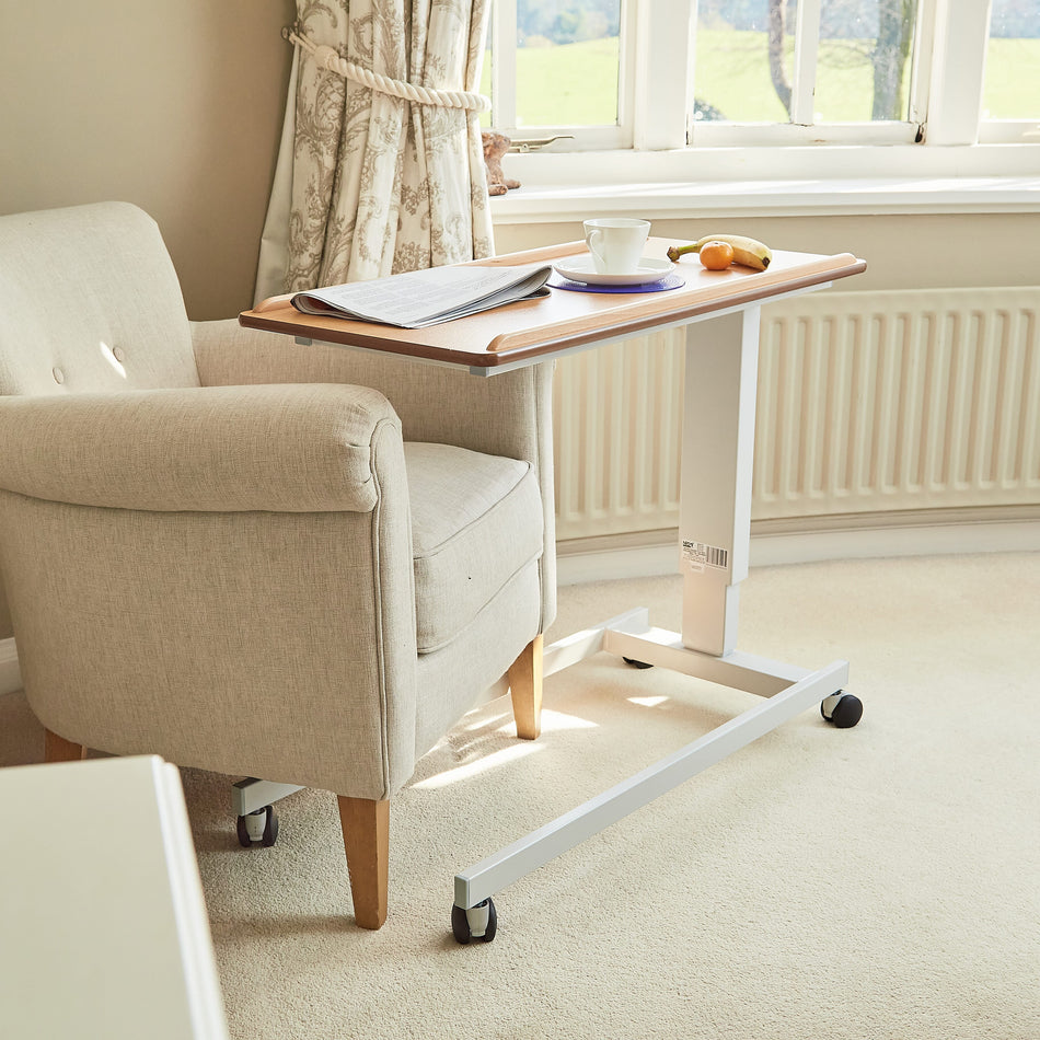 Angled view of NRS Easylift Overbed Table-Low with beech-effect tilting table top and white U-shaped base on castors against a neutral background.