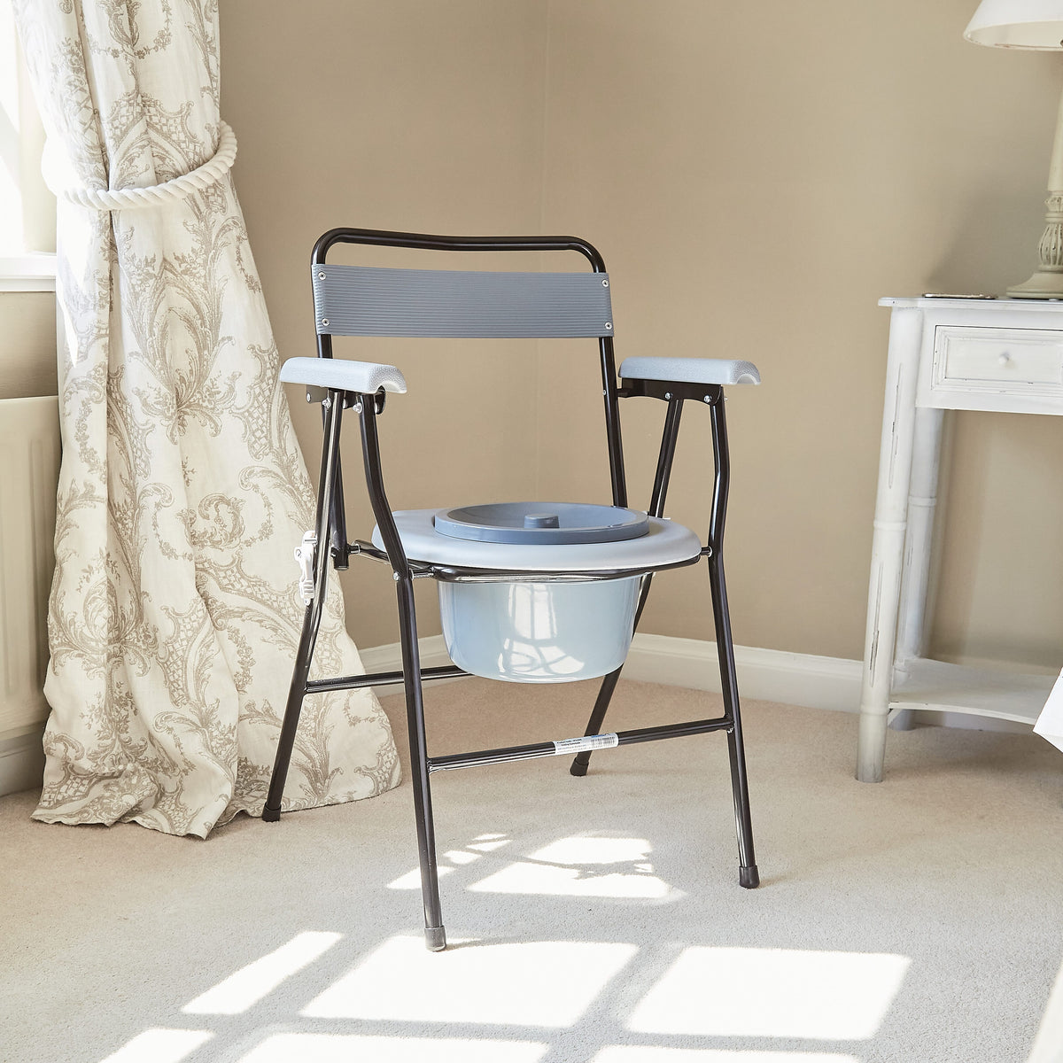 NRS Folding Commode with lid raised showing the open seat and removable commode pan, with supportive arms and adjustable height on a plain background.