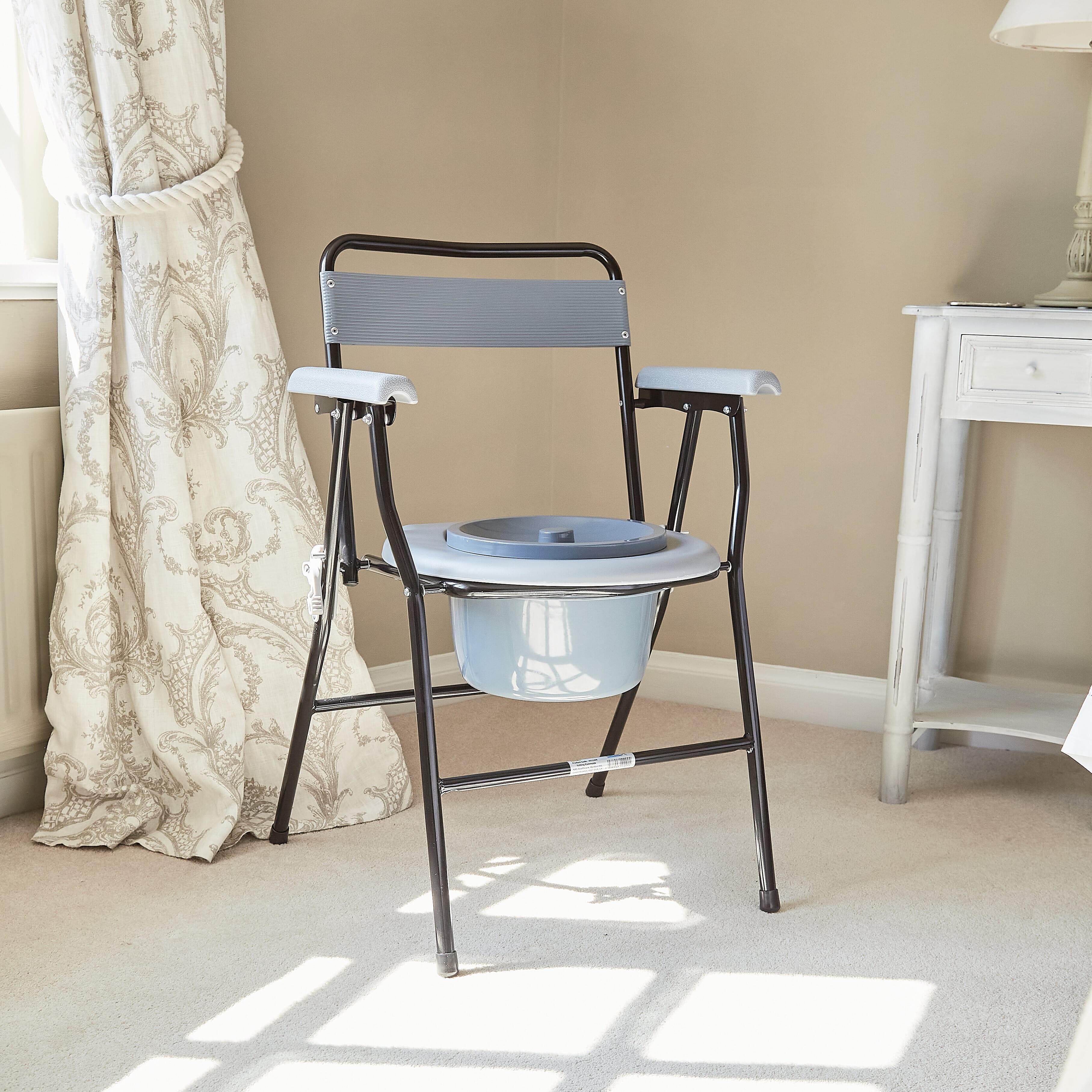 NRS Folding Commode with lid raised showing the open seat and removable commode pan, with supportive arms and adjustable height on a plain background.