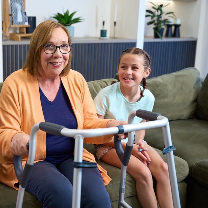 NRS Walk Safe Walking Frame helping an older adult stand up from a sofa with assistance from a friend or family member.
