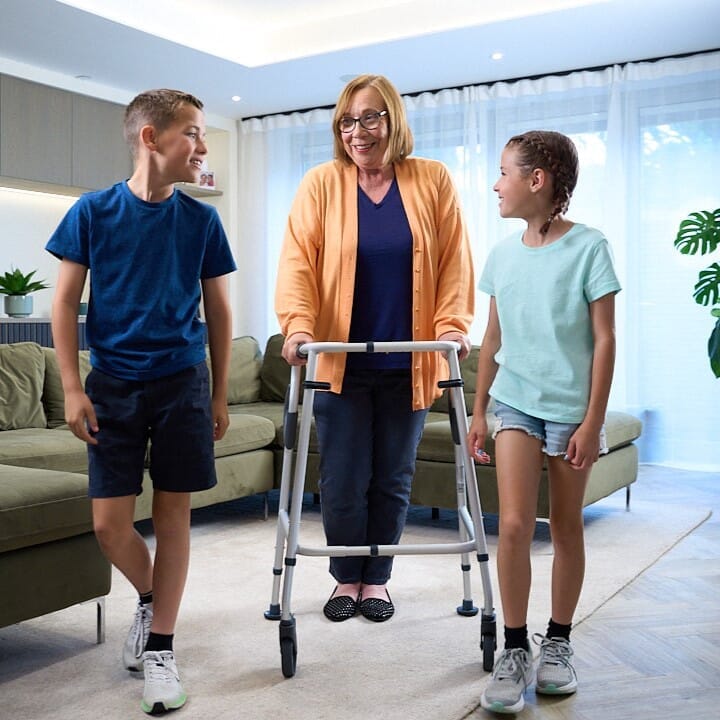 An older adult walking across a room with NRS Walk Safe Walking Frame accompanied by loved ones, illustrating mobility and independence.